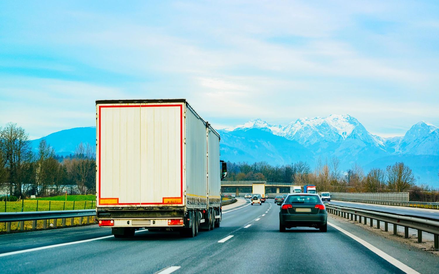 A large white freight truck driving on a highway with a car in front, against a backdrop of snow-capped mountains.