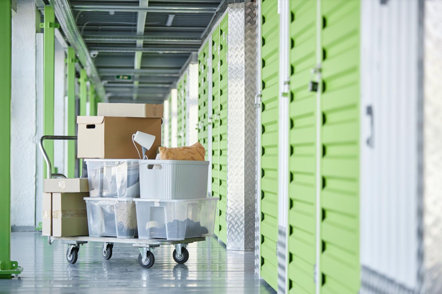 A metal cart loaded with cardboard boxes and clear plastic bins in a hallway lined with green self-storage unit doors.