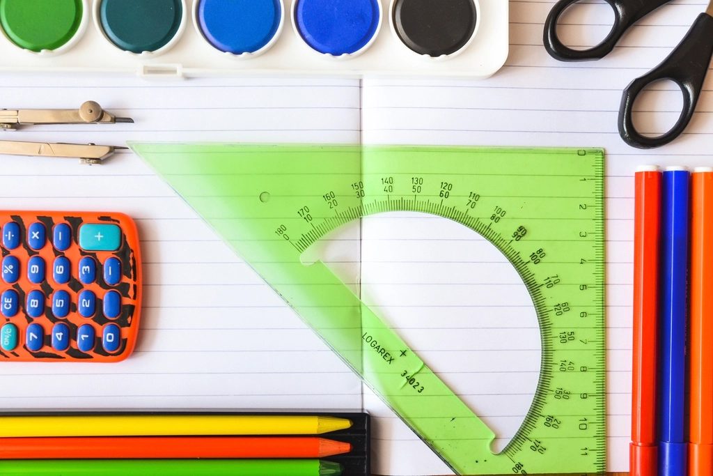 School supplies laid out on a desk, with a notebook, pencils, scissors, paints, and a ruler